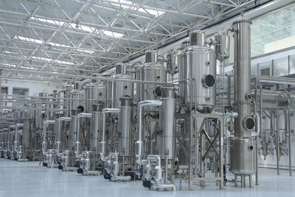 Multiple tall stainless steel evaporators and distillation columns with pumps and piping in a bright, clean industrial production hall