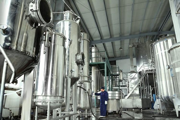 Worker in protective clothing inspecting tall stainless steel process tanks and piping inside a modern industrial processing facility
