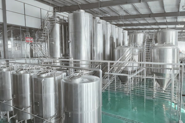 Multiple polished stainless steel storage and process tanks with platforms and stairs inside a bright, clean industrial production workshop