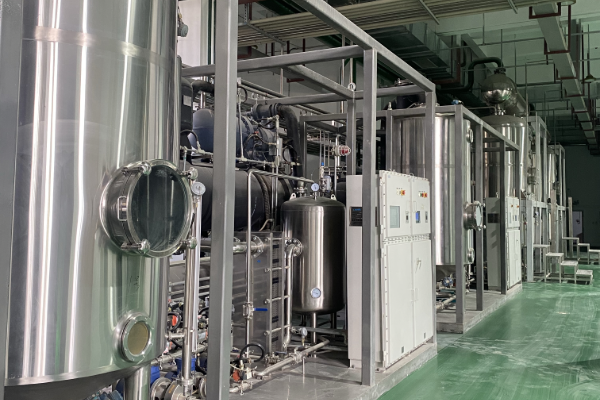 Row of skid-mounted stainless steel process units with columns, vessels, piping and control cabinets inside a clean industrial distillation workshop