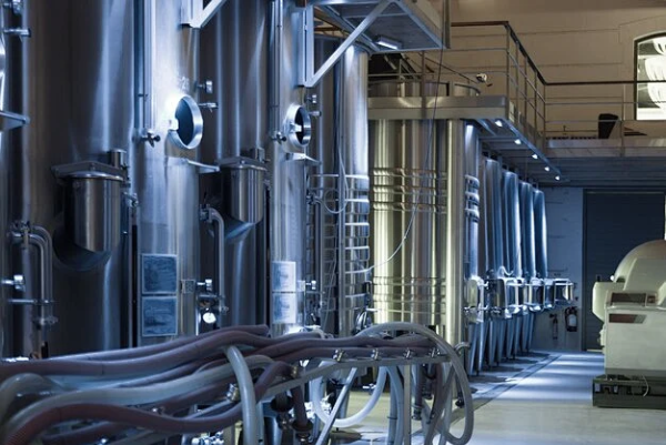 Row of tall stainless steel process tanks with hoses, piping and upper walkways inside a modern industrial production facility