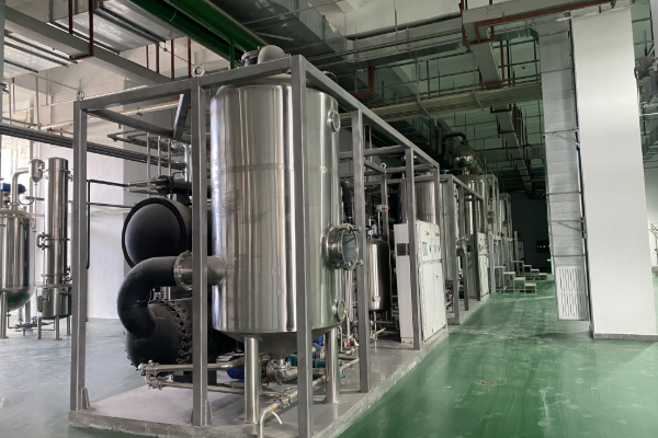 Row of skid-mounted stainless steel vessels, heat exchangers and control cabinets installed on a green-floor clean industrial processing workshop
