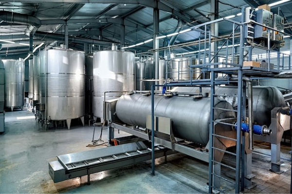 Industrial hall with multiple stainless steel storage tanks and a horizontal processing vessel with platforms, piping and conveyor inside a modern production facility