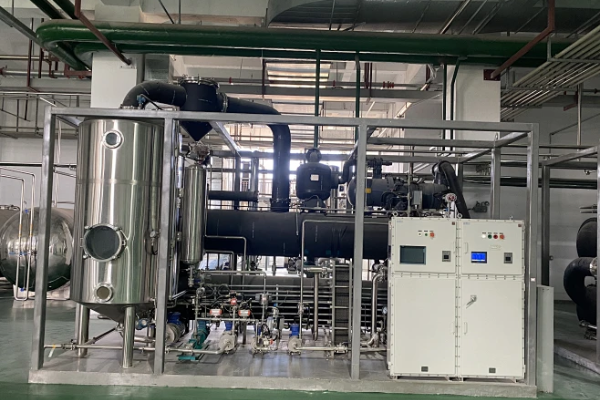 Skid-mounted stainless steel vessel, black compressor and heat exchanger unit with white control cabinets inside a clean industrial distillation workshop