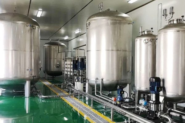 Row of stainless steel storage and process tanks with pumps and sanitary piping inside a bright, clean industrial process room
