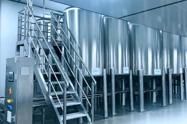 Row of tall stainless steel process tanks with conical bottoms, access stairway and sanitary piping inside a bright, clean industrial production room