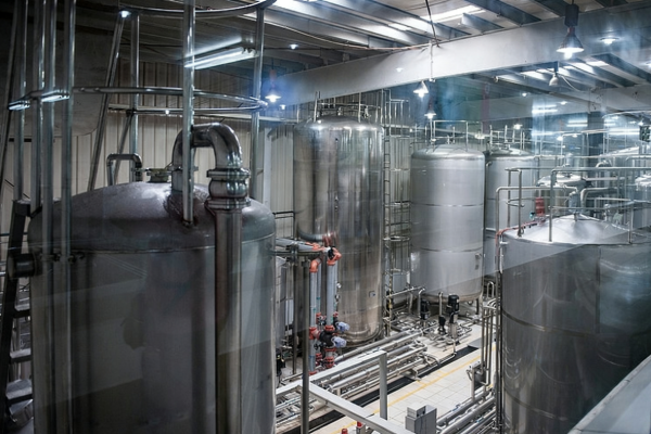 Multiple stainless steel process tanks and piping with overhead walkways and lighting inside a modern industrial production facility