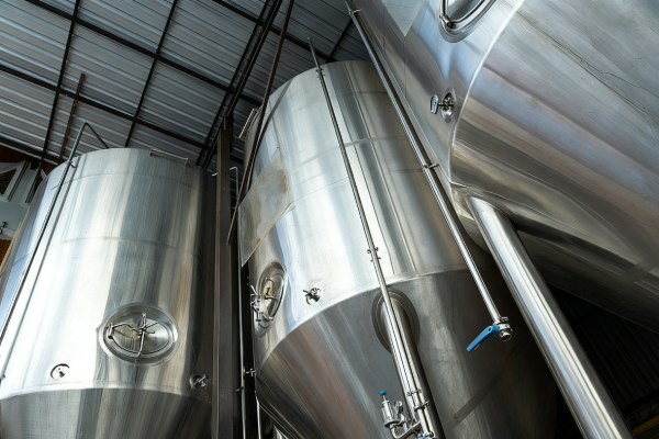 Upward view of large stainless steel storage tanks with piping and valves beneath a metal roof in an industrial production facility