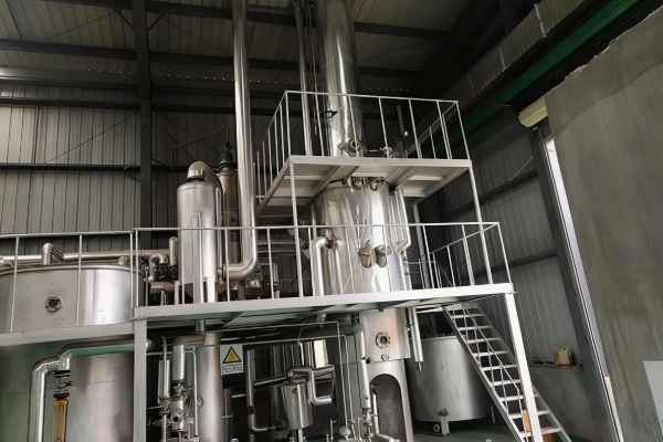 Tall stainless steel distillation column with platforms, piping and access stairs inside a modern industrial processing workshop